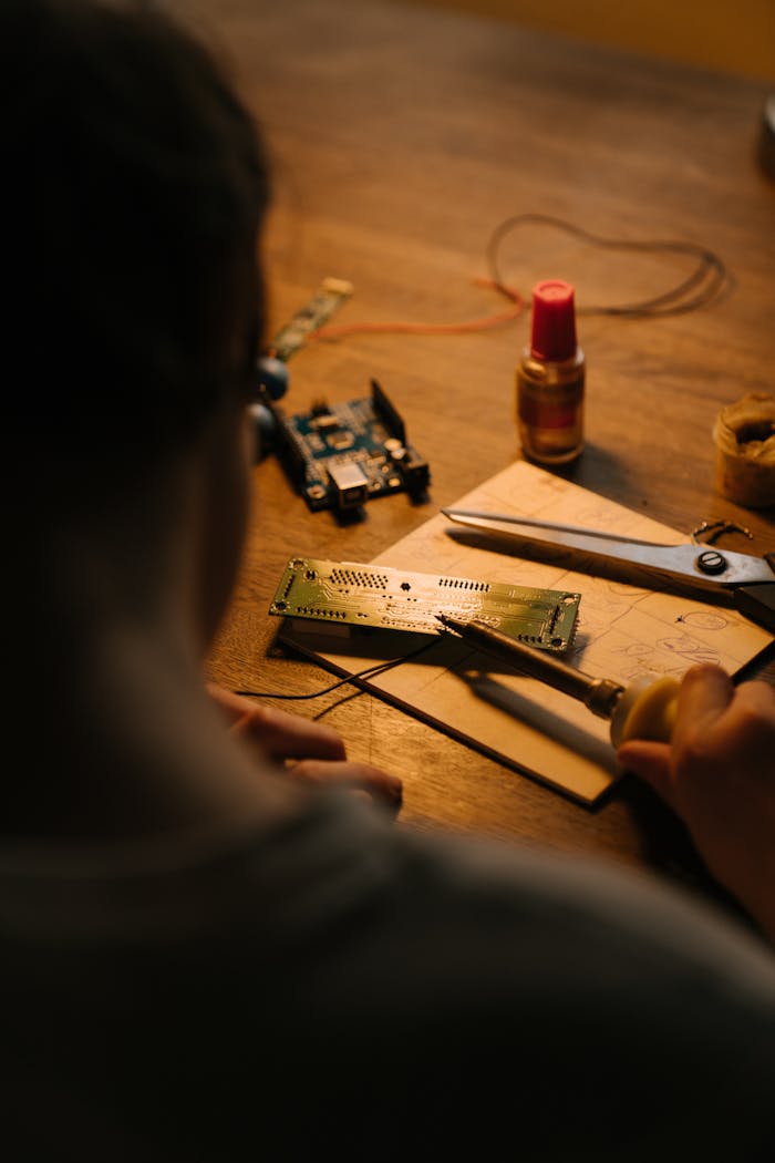 Person soldering a printed circuit board at a workstation with tools and components.